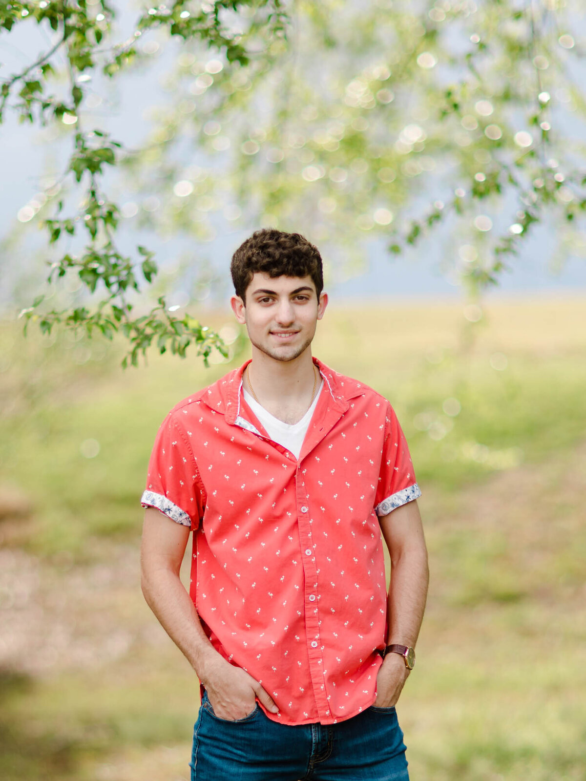 Senior portrait of a guy in an orange button down shirt standing in the middle of leaves from a tree and a blue sky behind him.