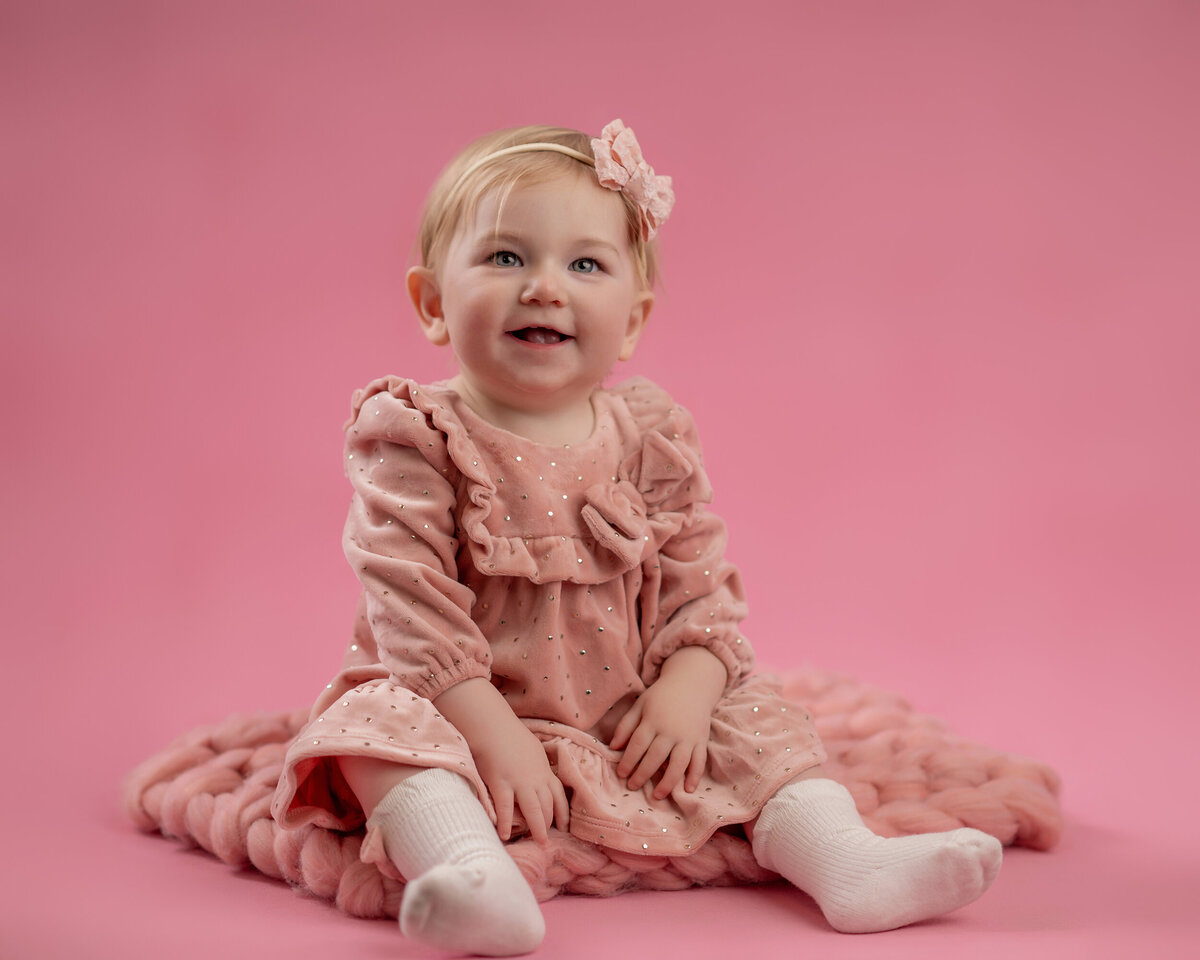 Wearing a pink dress, a young girl smiles while she sits on a pink background