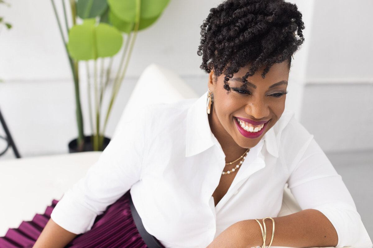 Discover the stunning portrait of a joyful African-American woman, captured in a light-filled Cincinnati studio. She radiates confidence and elegance, wearing a white blouse and a purple skirt, her natural curls perfectly styled. Ideal for personal branding and professional headshots, this image showcases the artistry of our Cincinnati-based portrait photography studio.