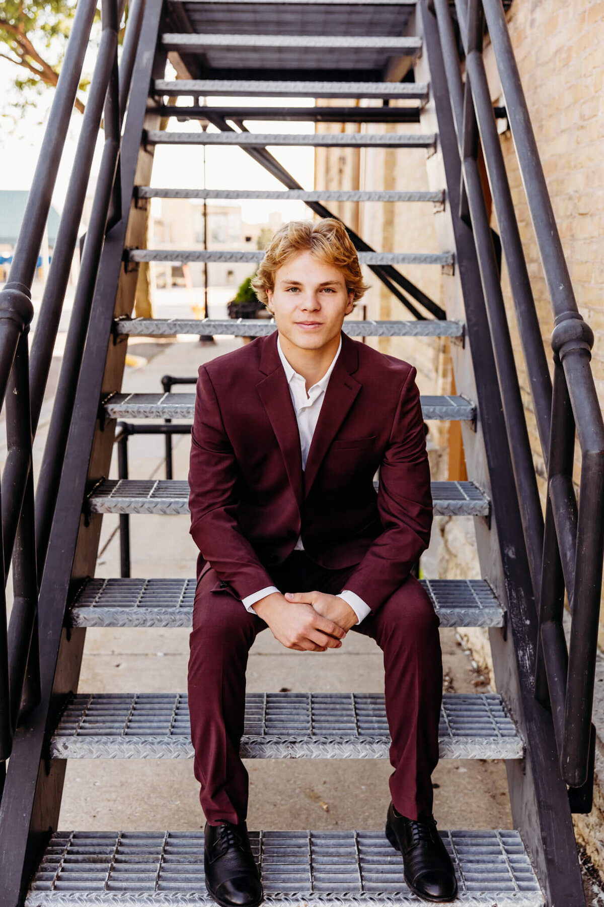 young man in a maroon suit sitting on a medal steps and holding his hands together downtown Neenah. 