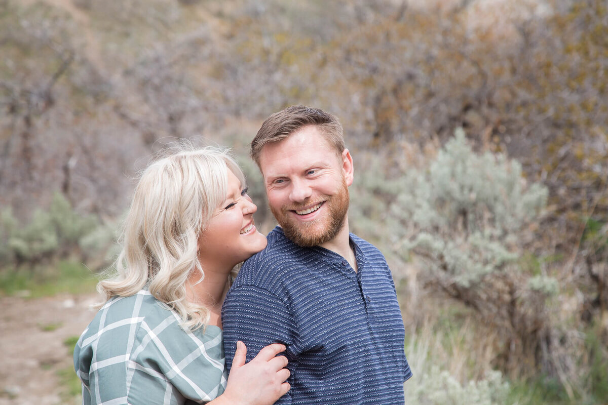 blonde wife smiling and snuggled up to bearded husband in blue shirt at mountain park