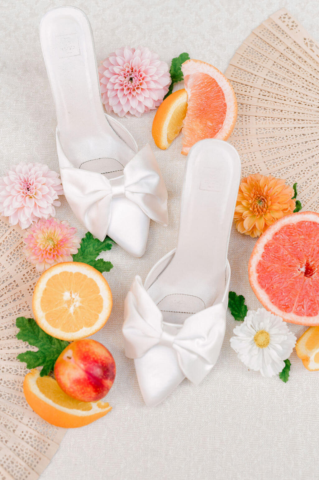 White bridal shoes with bows styled with citrus fruits and pastel flowers for wedding flatlay.