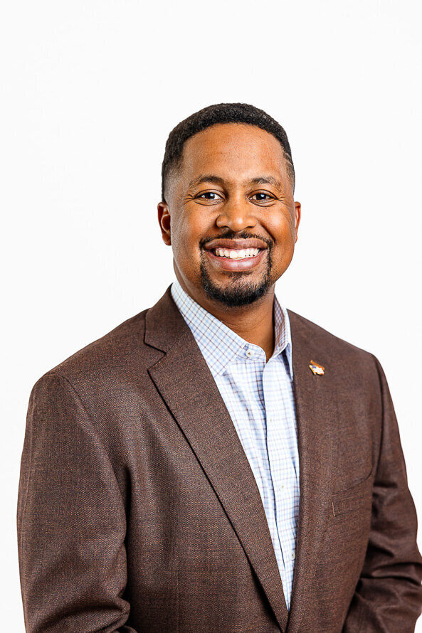 Man in brown jacket smiles for camera in a white photo studio.