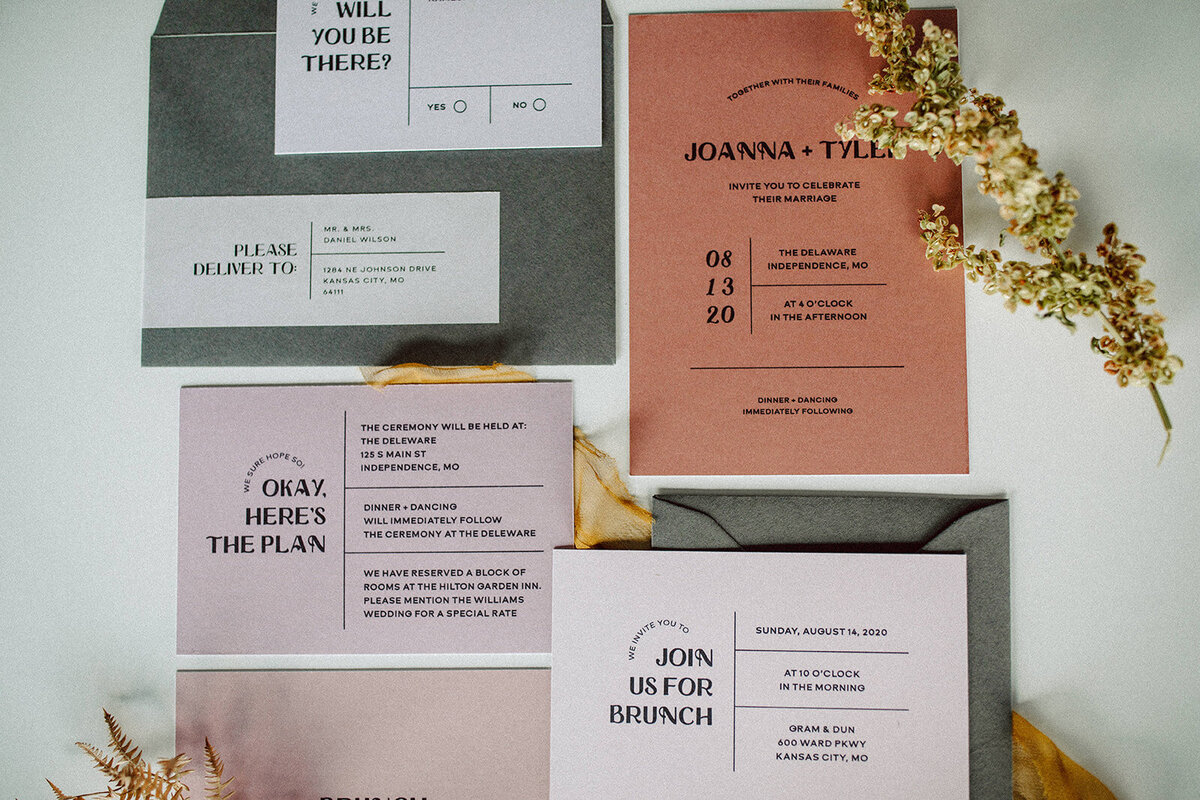 Peach, mauve, and off-white wedding stationery with black font atop a white table with stems of flowers.