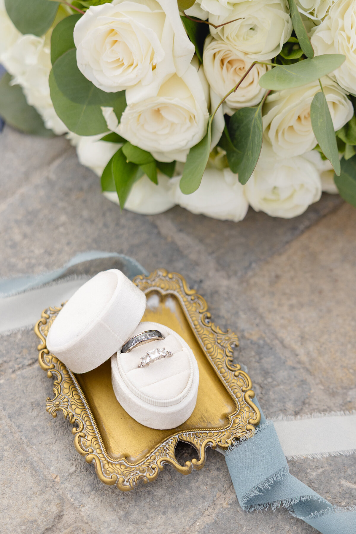 A close-up of a wedding ring set in a cream velvet box on an ornate gold tray, placed beside a bouquet of white roses and greenery on a stone surface.