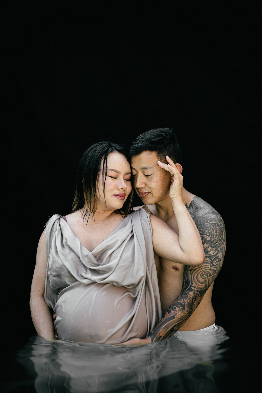 Pregnant woman stands in a pool with her partner and they both hold her bump