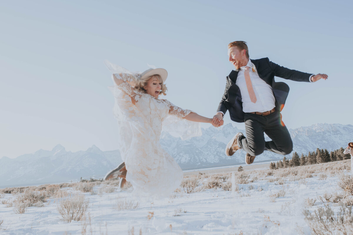 Idaho Falls wedding photographer documents winter elopement with bride and groom holding hands and jumping in the air while the bride holds her hat