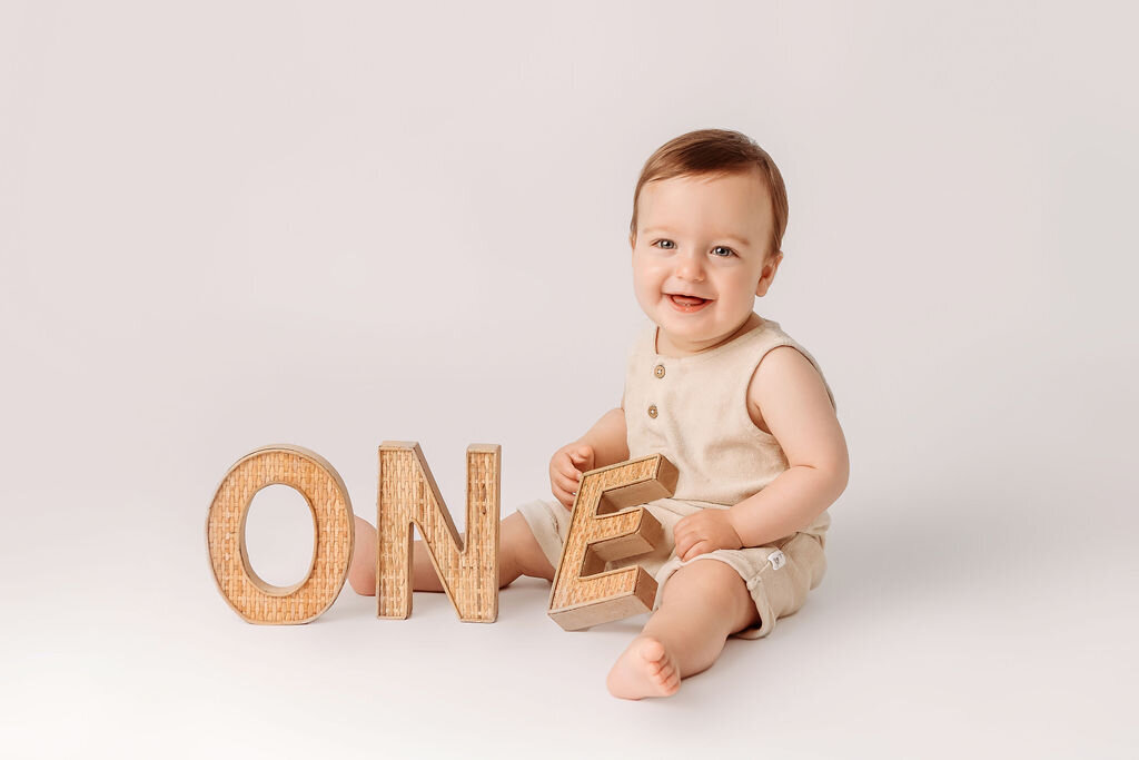 Portrait Photographers CT capture smiling baby boy in neutral linen outfit sitting with large wooden ONE letters during minimalist first birthday portrait session