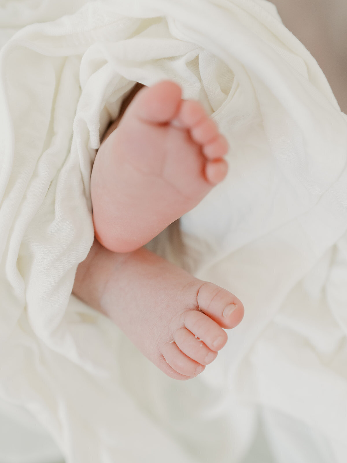 A close-up photo of a newborn’s tiny feet, curled slightly and resting on a soft blanket. The delicate wrinkles and tiny toes are in sharp focus, highlighting the baby's innocence and newness. The warm, natural lighting adds to the softness and tenderness of the image.