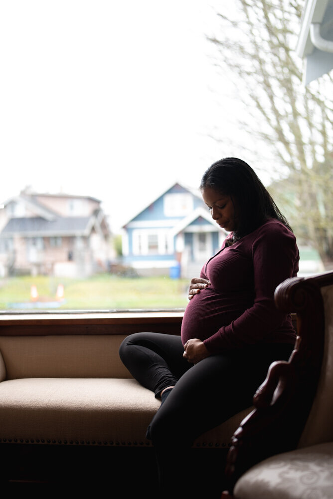 Mom wearing comfortable clothers sitting in a window seat of her home, cradling her pregnant belly