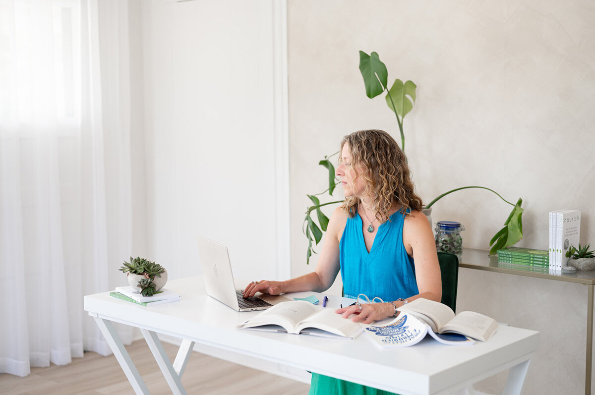 Nutritionist Supervisor, Heidi Schauster works at a bright, modern desk with an open laptop and books. She is surrounded by plants, creating a calm, focused atmosphere.