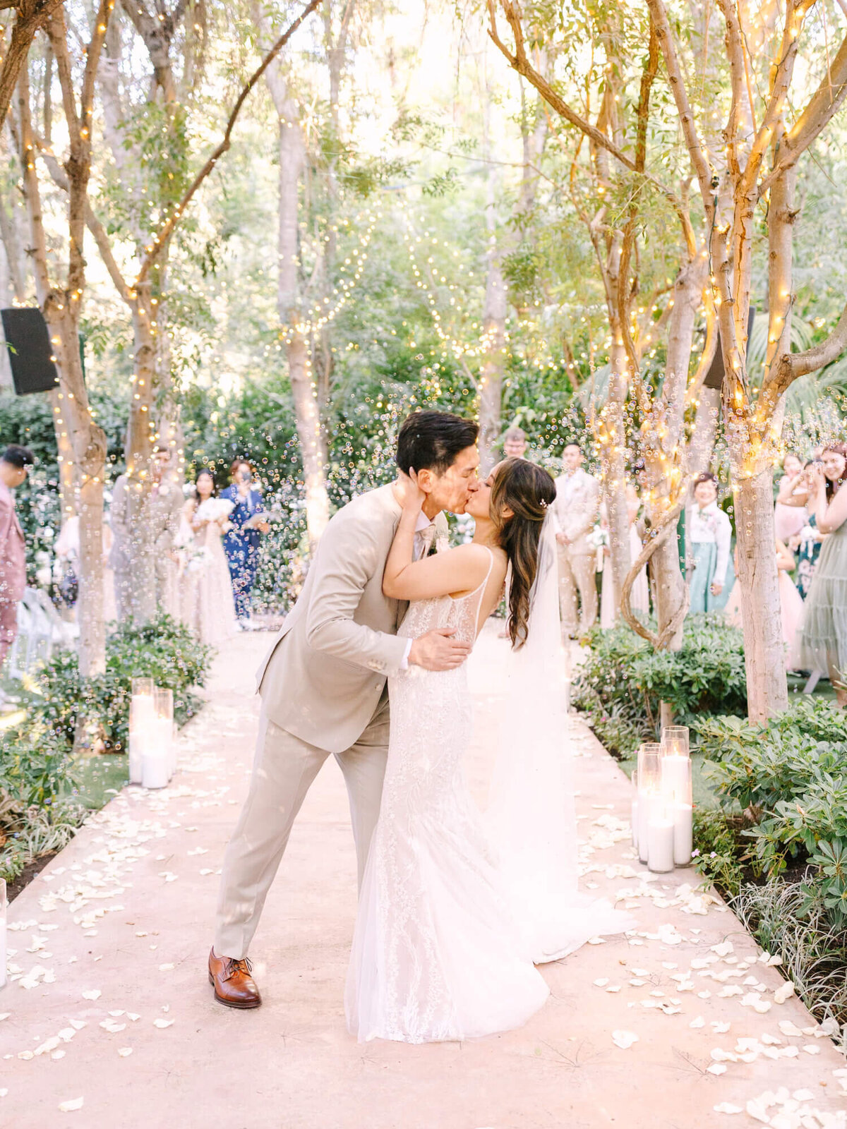 A couple kisses under twinkling lights and trees in a lush garden setting. Guests watch happily in the background.