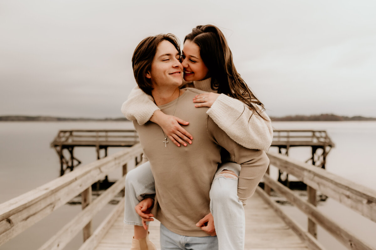 outdoor engagement session with woman piggy backing on her fiance as they are on a dock in the lake and he looks back at her for their little rock engagement photos