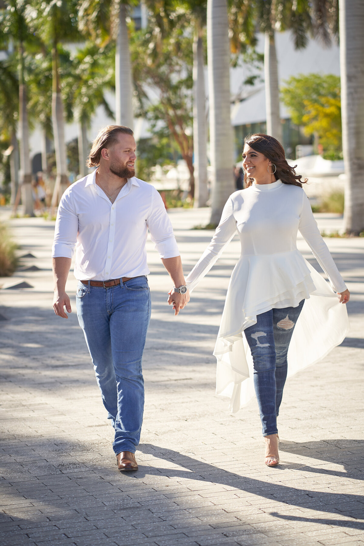Couple walking hand-in-hand in a sunny Miami park, smiling at each other, surrounded by palm trees and warm natural light.