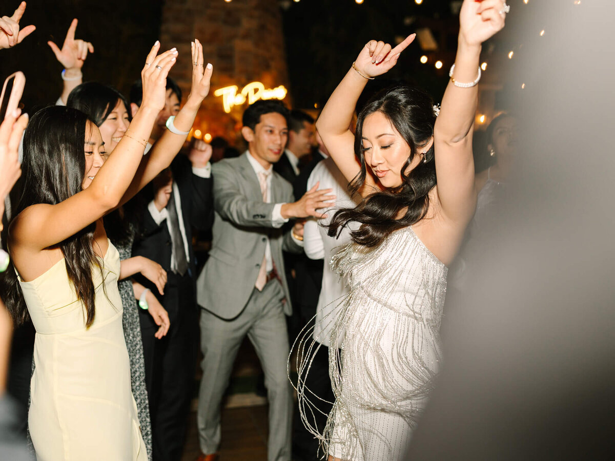 A joyful group of people dancing at an event, with a bride in a shimmering dress raising her arms. Warm lights and festive atmosphere in the background.
