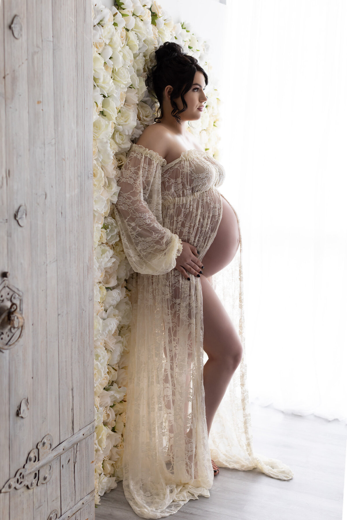 A serene maternity portrait featuring a woman gently resting against a lavish white rose wall, wearing an ethereal lace gown. The soft natural light highlights her tranquil expression and elegance, captured by Lindsay Walden Photography.