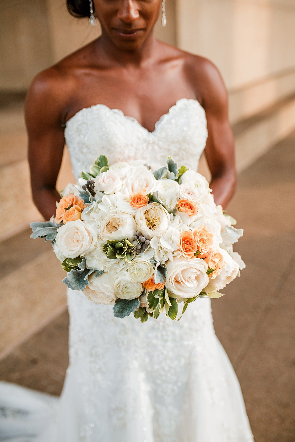 An African American bride wearing a strapless mermaid wedding dress with beaded lace holds a large round bridal bouquet in front of the Parthenon in Nashville. The bouquet has ivory peonies, white roses, peach tea roses and orange roses with white tipped greenery.