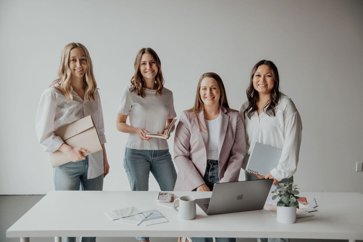Four women smiling holding folders, books and a laptop over a desk