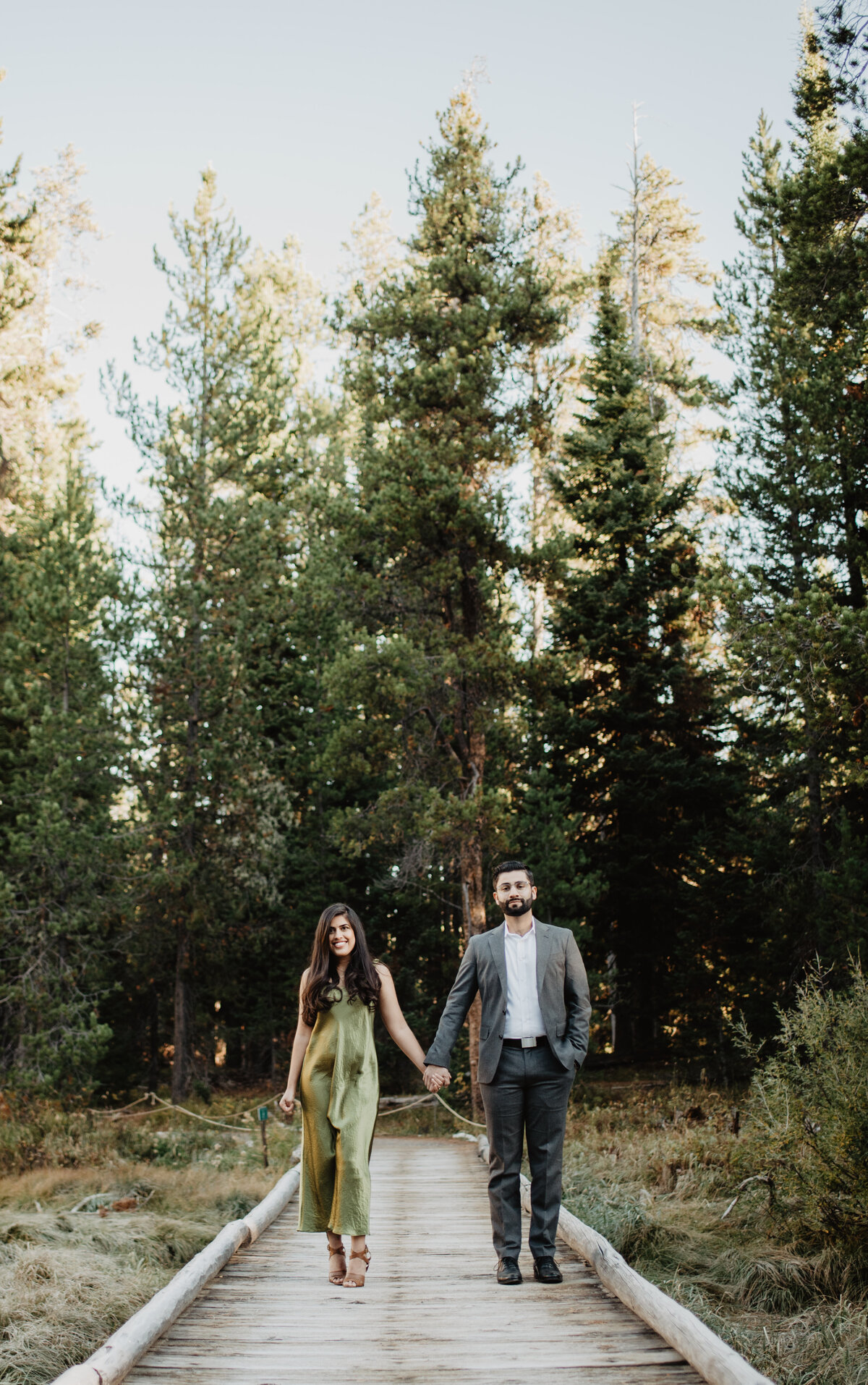 man and woman holding hands and walking on a wooden dock towards the camera with a forest behind them for their fall engagement session in Jackson Hole