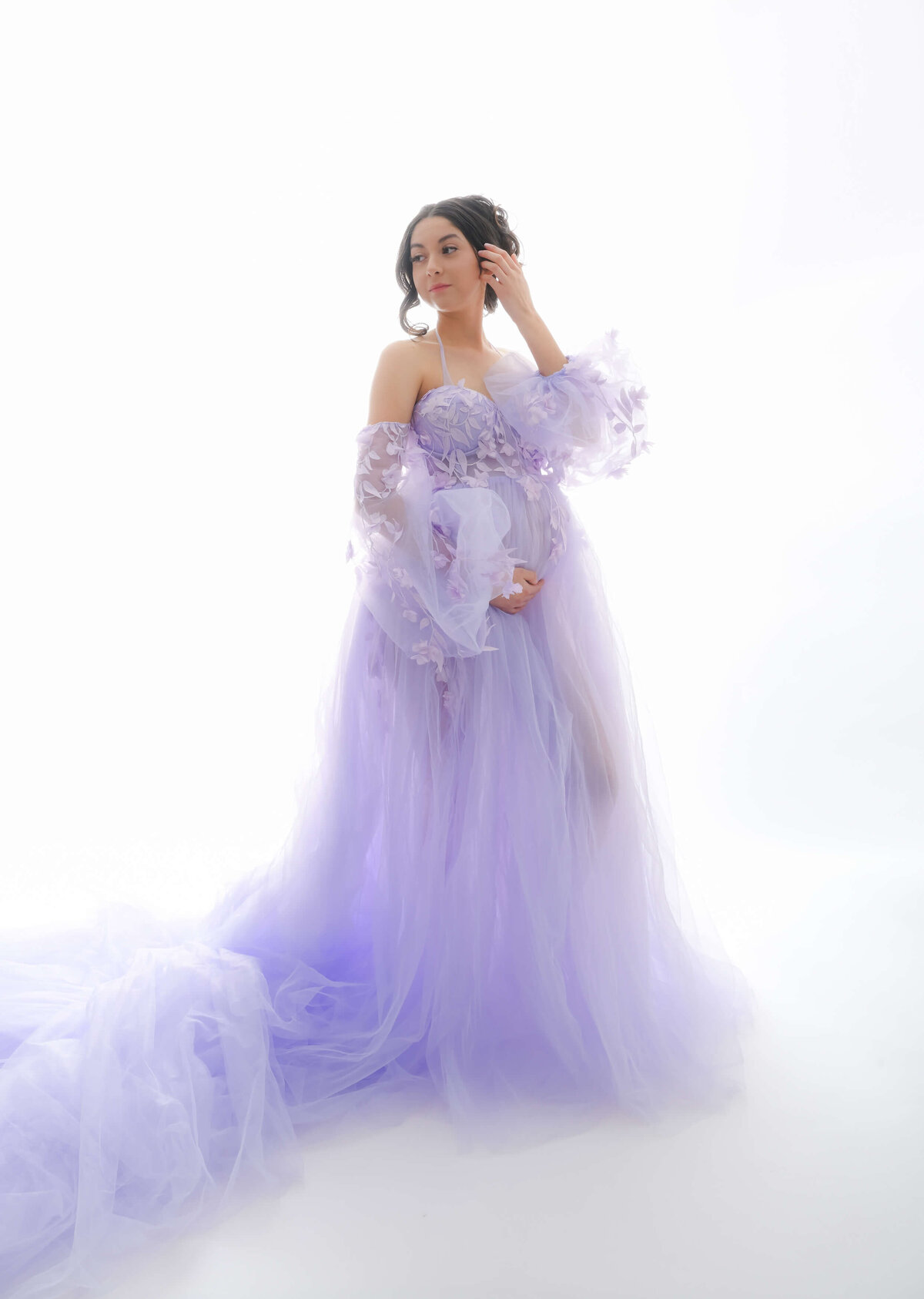 Maternity photo session with back lighting.  E. Benecki Photography captures expectant mother looking over her shoulder with one hand gently placing hair behind ear and other hand cradles her baby bump. She is wearing a floor length, lavender, off the should dress.