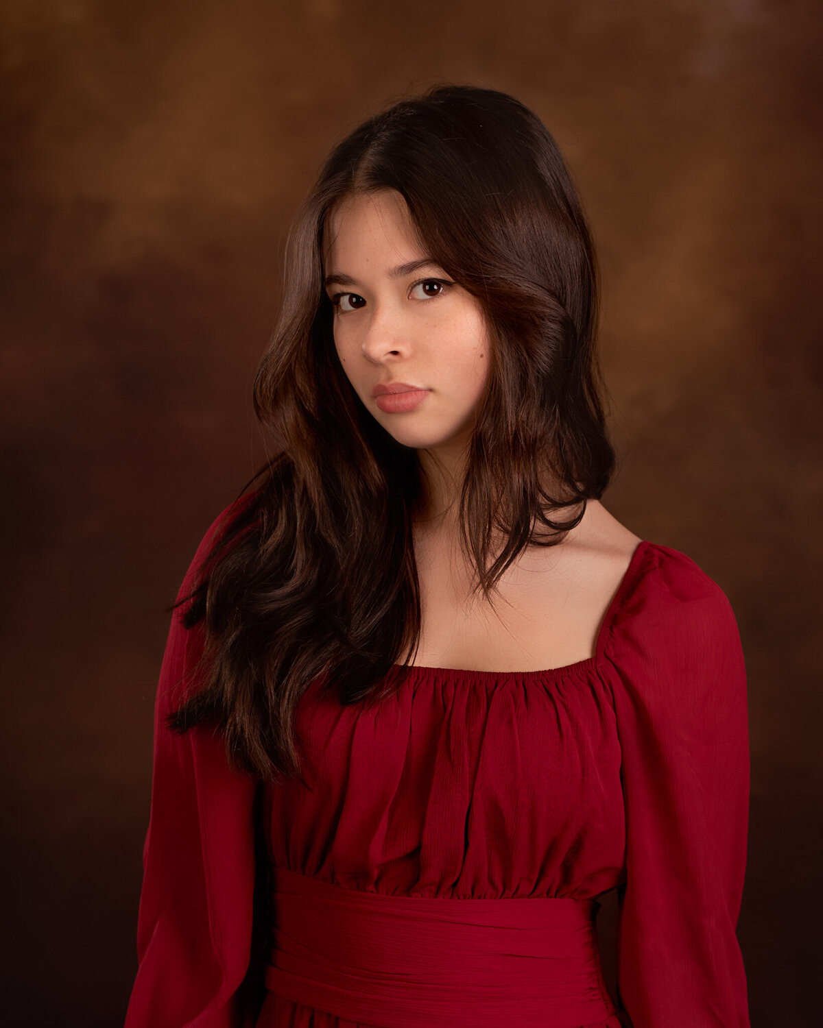 Elegant fine art portrait of a young girl in a flowing red gown, posed against a rich brown backdrop. Captured in Overland Park, Kansas, this classic and dramatic setup is ideal for families seeking polished and artistic child photography