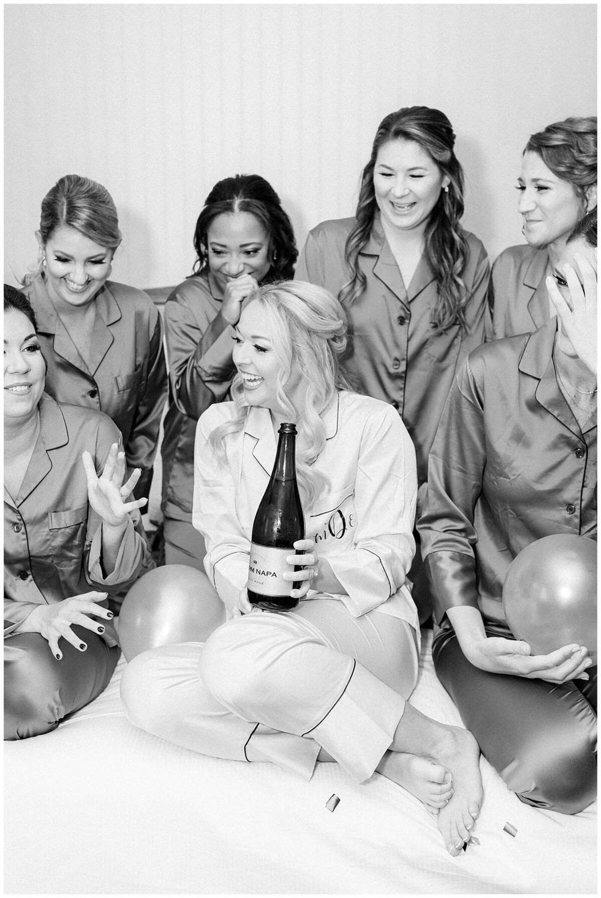 Black and white photo of bride holding champagne and laughing with her bridesmaids around her