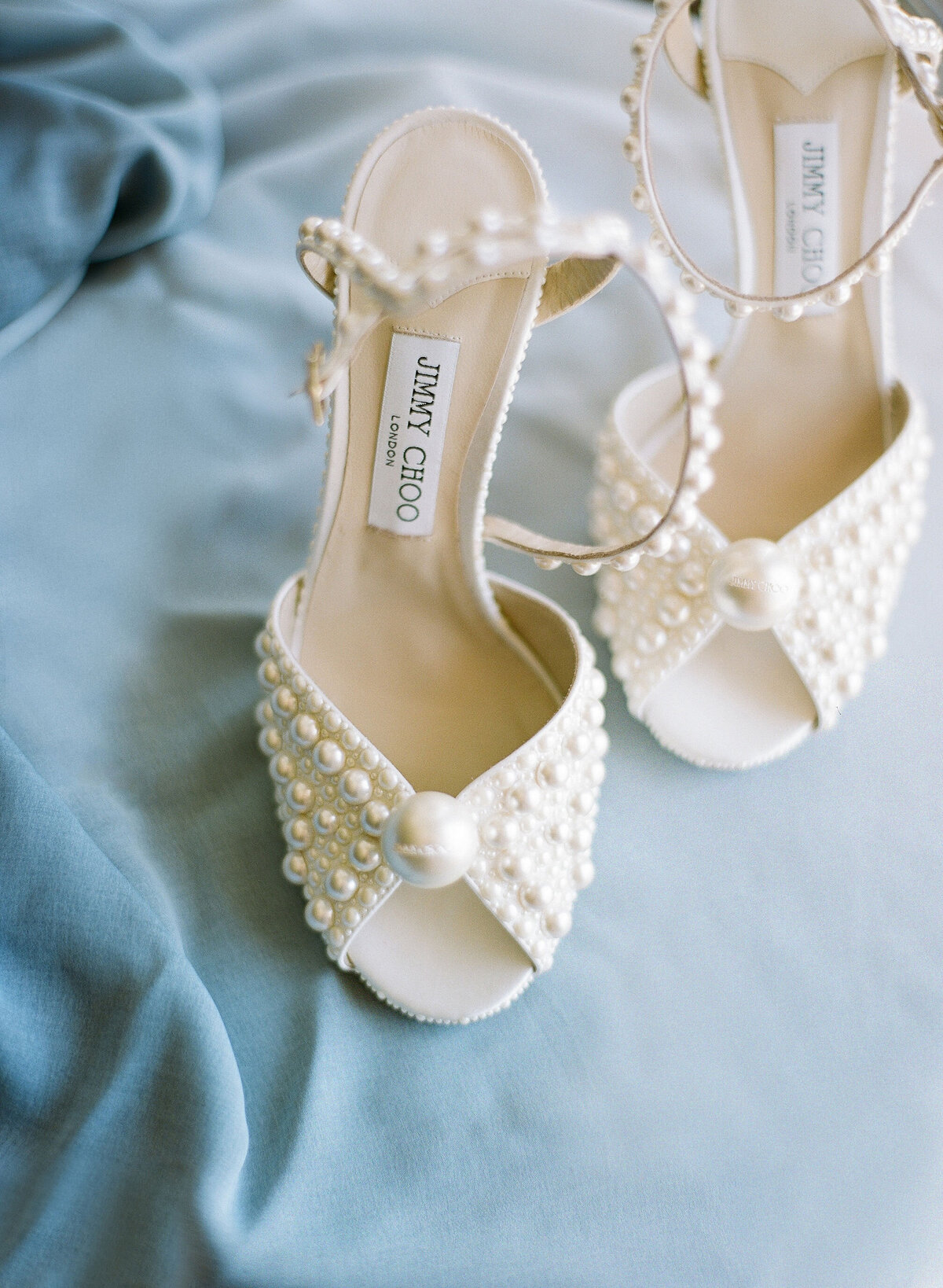 The photo was taken at Washington D.C., Washington National Cathedral by photographer Twah Dougherty. The image captures a pair of elegant open-toe high heel shoes adorned with pearls and a large pearl on each toe, placed on a soft, light blue fabric. The insole features the designer label Jimmy Choo, emphasizing the luxurious and stylish design of the shoes.