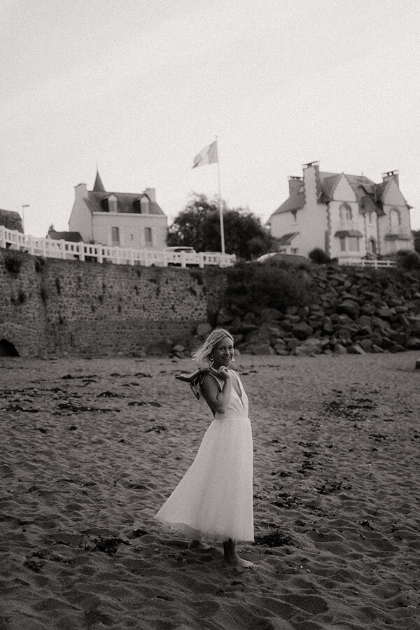 Portrait naturel de la mariée seule sur la plage, pieds nus et chaussures par dessus les épaules. Moment spontané par kamescop.