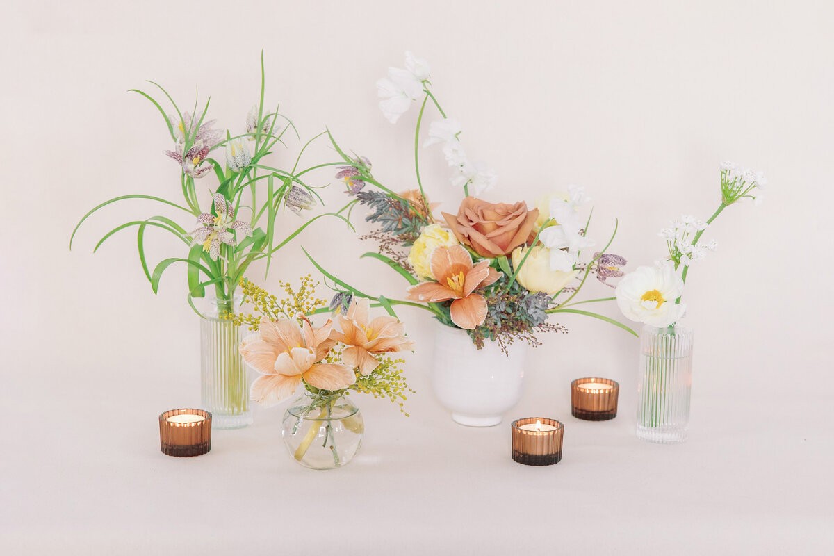 Four bouquets in vases are set against a white backdrop. Three candles surround them.