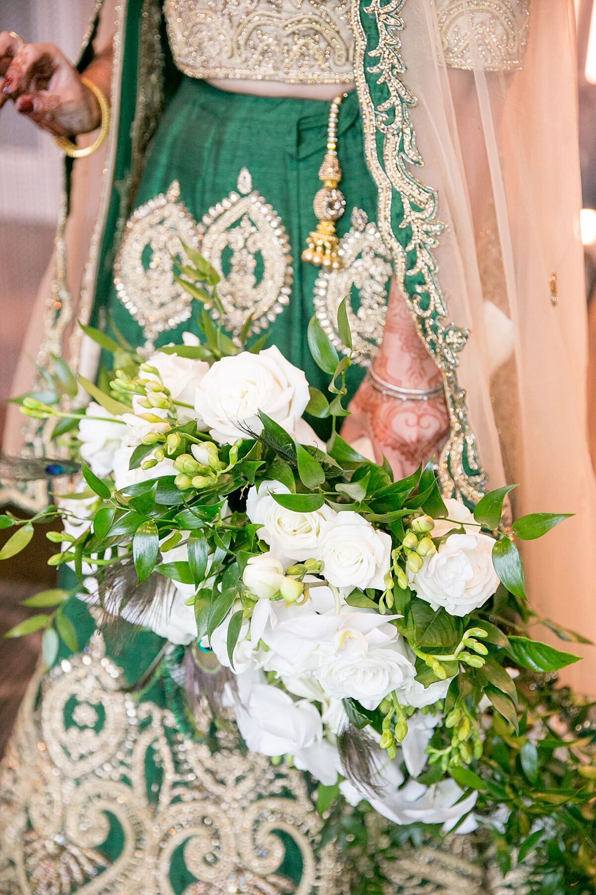 Indian bride wearing a gold and green saree holds a white cascading bouquet or orchids and roses as she displays her mendhi.