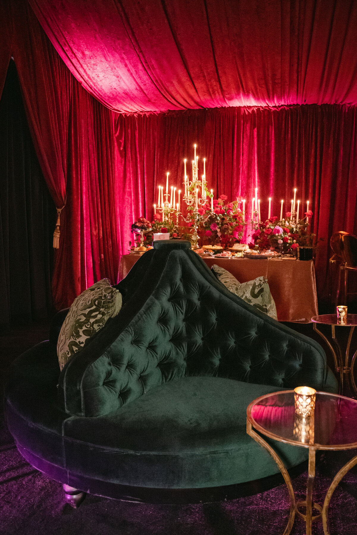 The stunning photo was captured at a tented wedding in Virginia by photographer Elisabetta Marzetti with videographers Clayton Films. The scene showcases a luxurious lounge area, complete with a plush dark green velvet sofa that exudes elegance. Surrounding the sofa are exquisite gold candlelit tables adorned with intricate designs, creating a sophisticated ambiance. Rich red velvet drapes drape the room, adding a touch of opulence, while an extravagant candlelit dining table stands as a captivating focal point against the backdrop.