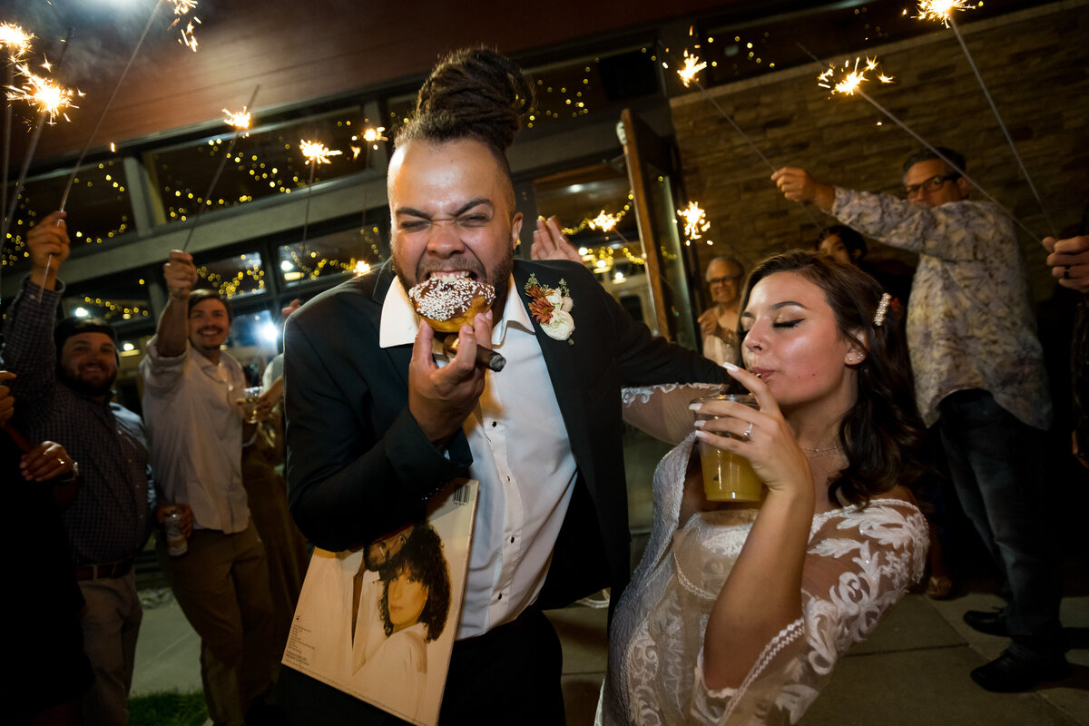 A bride and groom exit their reception at the Coeur d'Alene Casino with a drink and a donut while surrounded by family and friends.