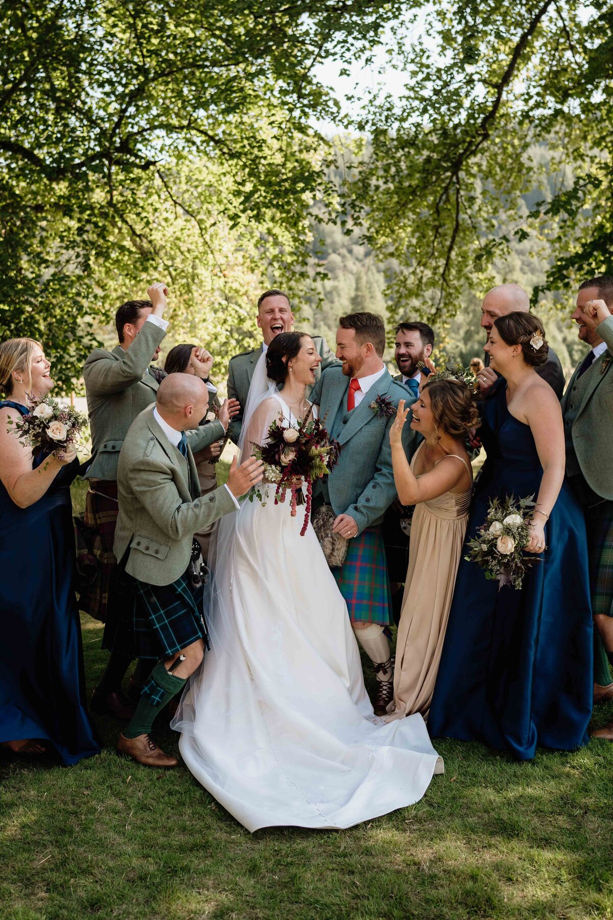 A wide photograph of a wedding party dancing around a bride and groom outdoors under trees at Mar Lodge.