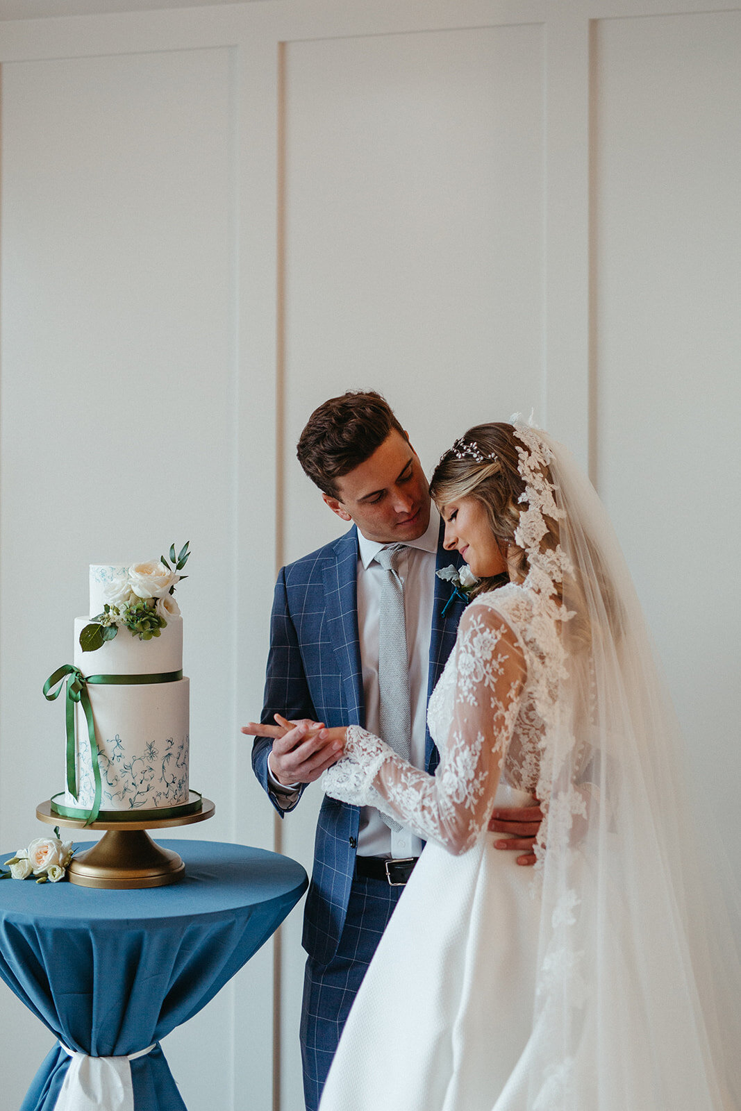 Bride and groom in a blue suit and white wedding gown next to a layered cake on gold cake stand atop a round table.