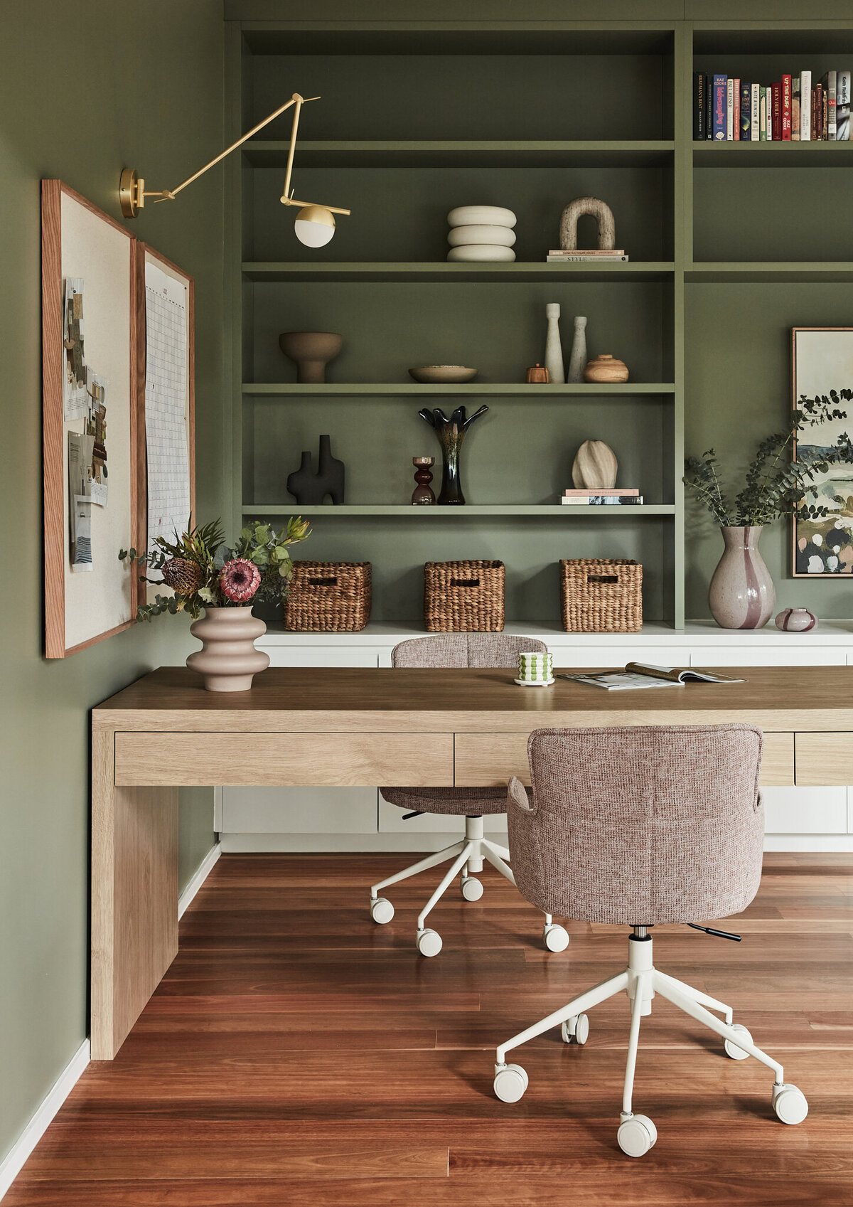 Detail of olive-toned office with wood desk, shelves styled with ceramics and baskets, and wall-mounted brass lighting in a Brighton home styled by interior designer Lisa Hunter.