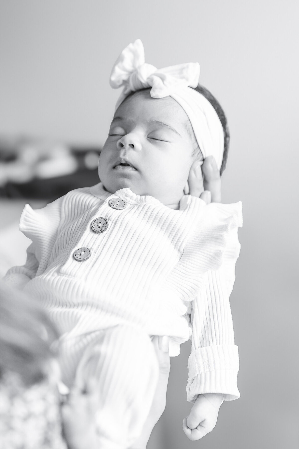 Peaceful newborn baby girl dressed in a ribbed ivory outfit with wooden buttons and a soft headband bow, cradled in her mother’s hands. A serene moment from an in-home newborn photography session capturing the beauty of early days.