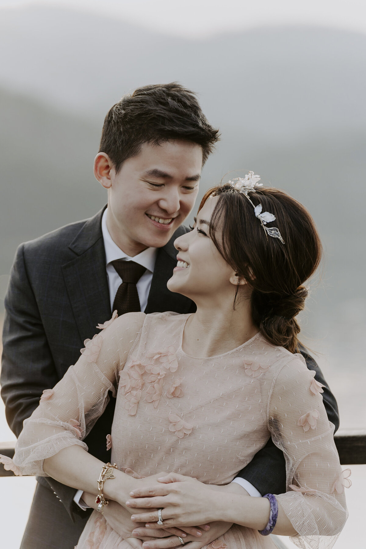 A groom wearing a suit hugs a bride wearing  a pink floral dress from behind