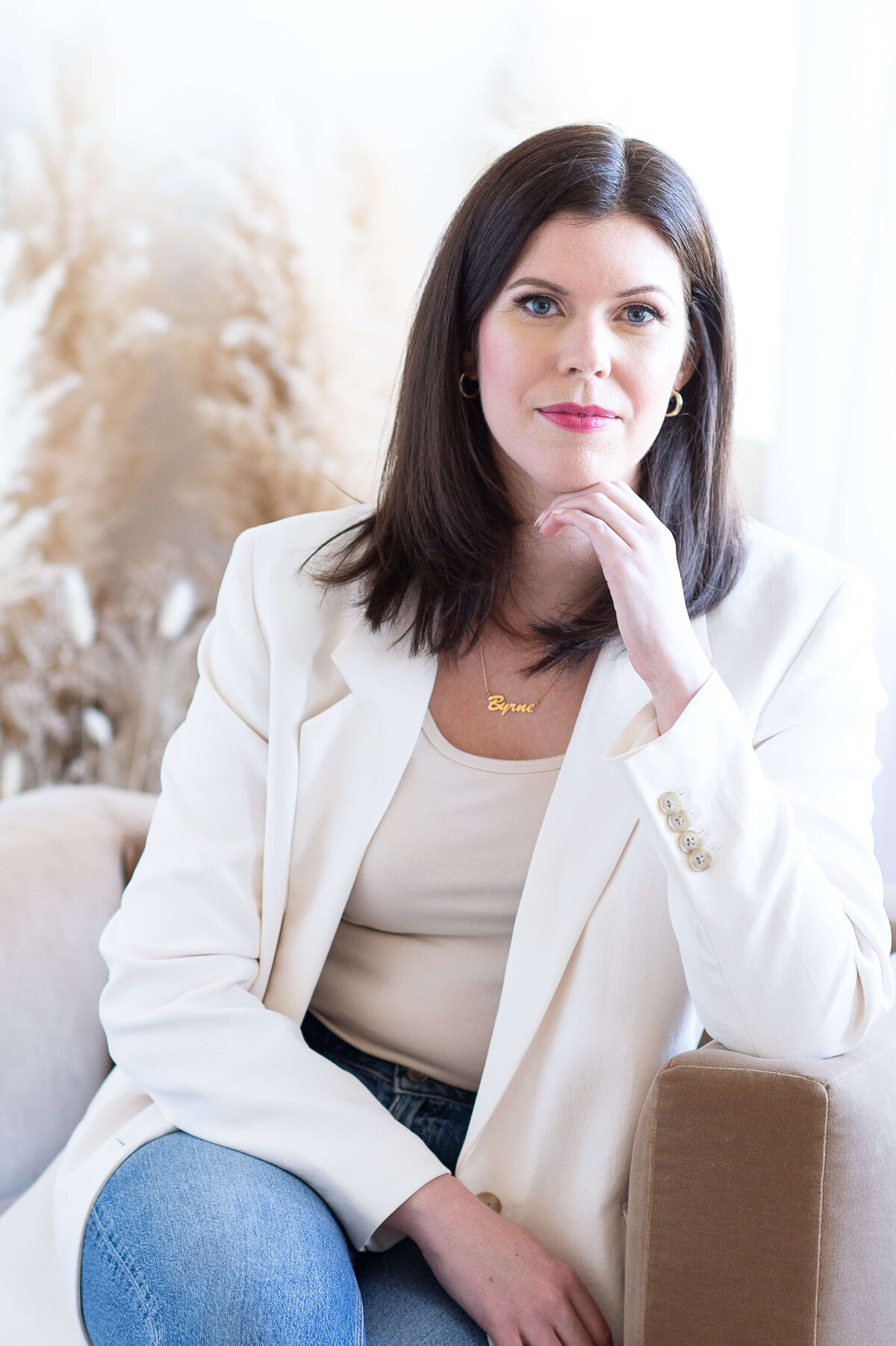 a woman in a white blazer sitting in a camel coloured chair with a pensive look on her face.  Captured indoors at Skye Studio by Ottawa branding photographer JEMMAN Photography