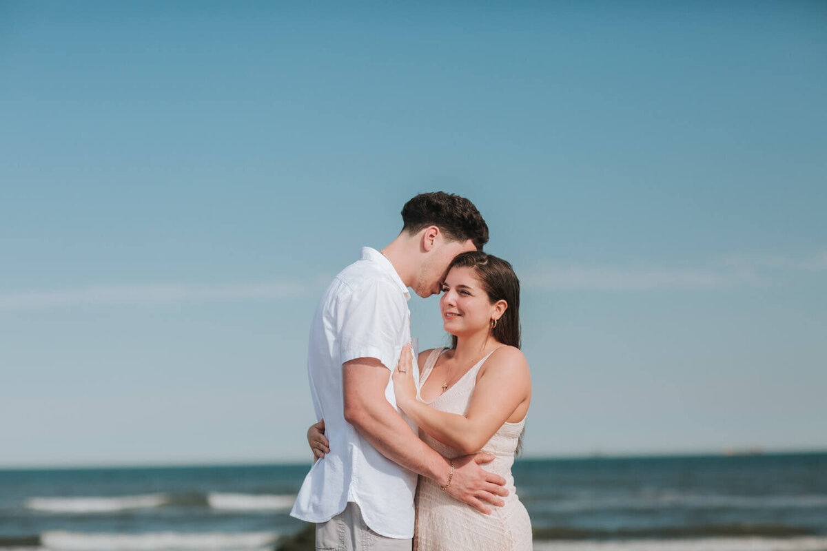 A couple embraces on the beach, with the ocean waves and clear blue sky behind them, at Wildwood Beach in NJ.