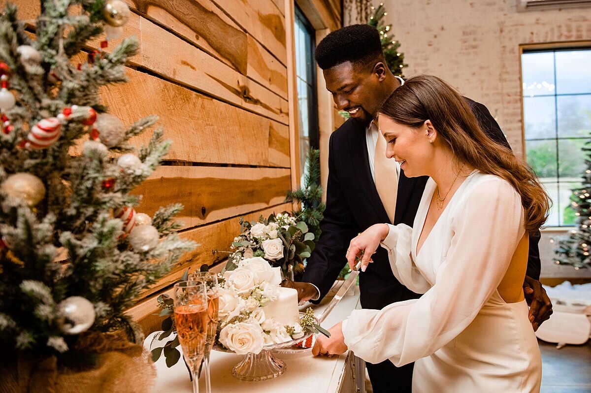 The bride, wearing a backless satin wedding dress with long sheer sleeves cuts the wedding cake with the groom who is wearing a black suit with a white shirt and gold tie. The cake is on a footed crystal cake stand and is decorated with white and blue flowers with eucalyptus. They are facing a cedar wood wall with a white washed brick wall behind them.