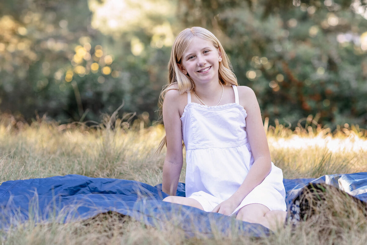 A young girl with blonde hair, wearing a white dress sitting on a blue picnic blanket in the park surrounded by greenery.