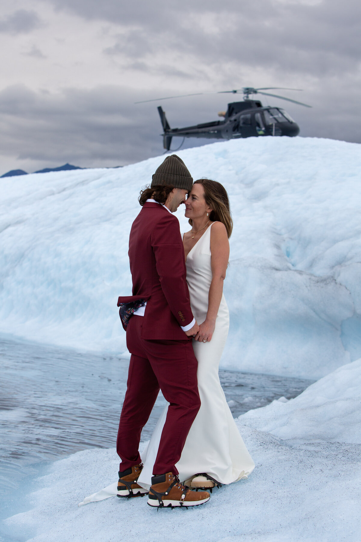 A bride and groom stand with their foreheads touching and holding hands on a glacier in Alaska with a helicopter parked on the ice behind them.