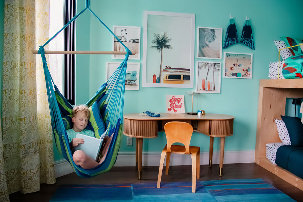 A young boy with short blond hair is comfortably seated in a blue and green striped hammock chair, reading a book. The hammock hangs near a window with patterned curtains in a bright room with light turquoise walls. Behind him, a light-colored Umage Audacious desk with tambour doors and slender legs stands against a gallery wall of framed beach-themed prints. A small lamp and a postcard reading "Stay Rad" are on the desk. A simple wooden chair is tucked underneath. To the right, a bunk bed with colorful bedding and a textured throw is partially visible. A colorful striped rug lies on the dark wooden floor.