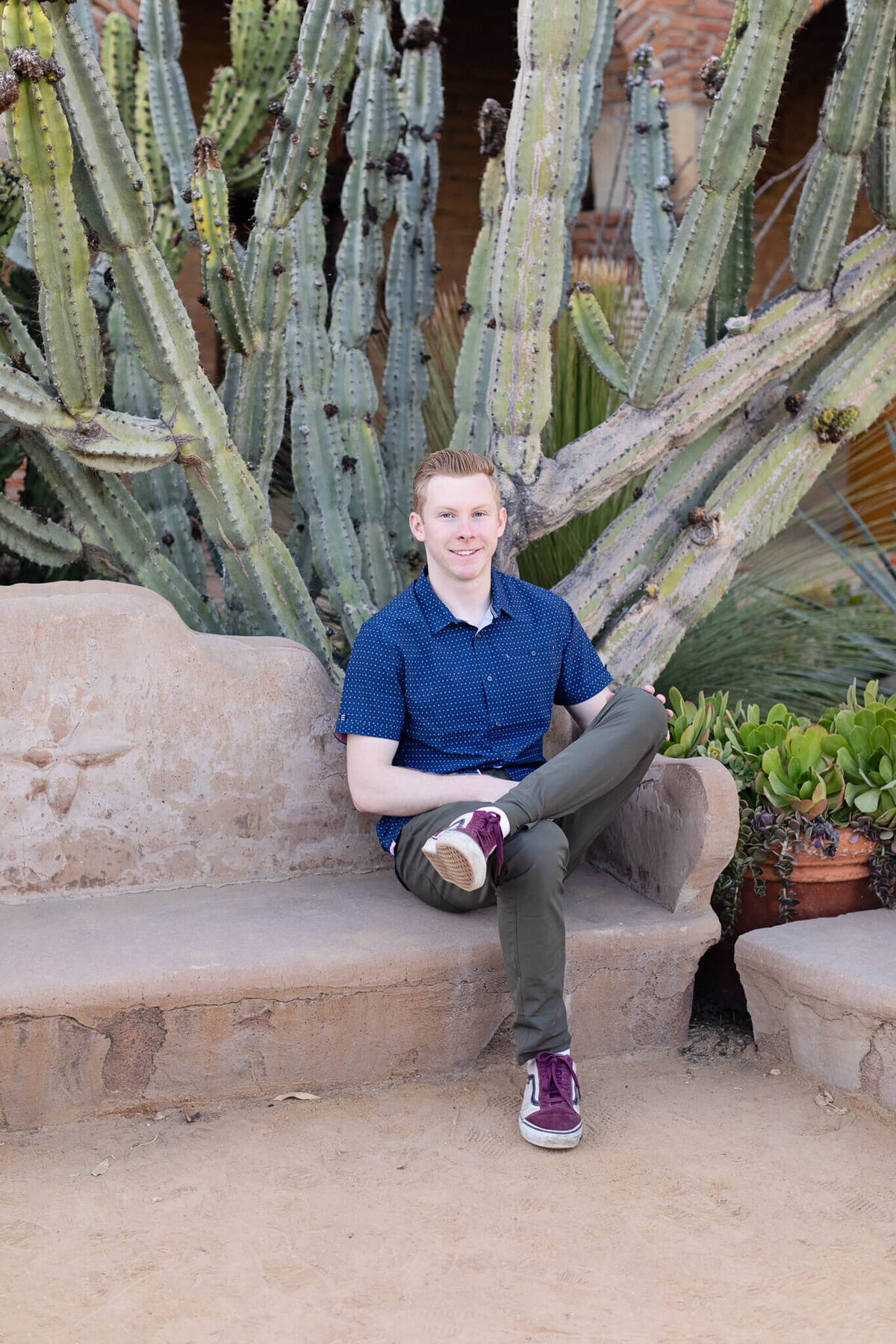 A young man with strawberry blonde hair sitting with one leg over the other on a light brown, clay bench in a blue button-up shirt, smiling at the camera. Captured at Mission San Juan Capistrano.