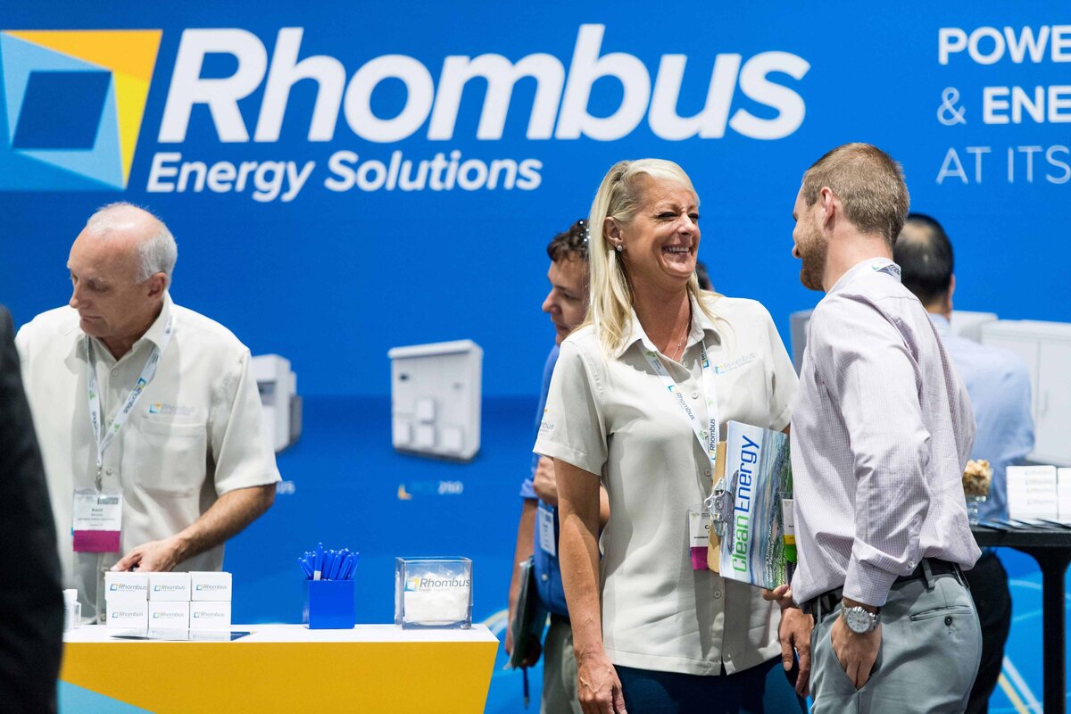 two adults interact at an energy solutions booth while a demonstration takes place in the background