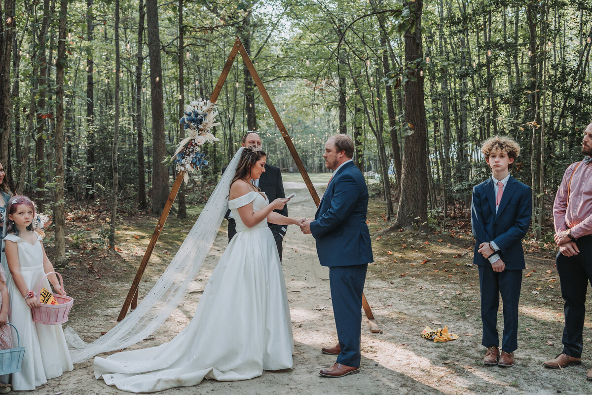A wedding couple exchanges vows under a rustic wooden triangle arch adorned with flowers in a beautiful forest wedding ceremony, surrounded by tall trees and loving guests, captured by Quinn Photography LLC.