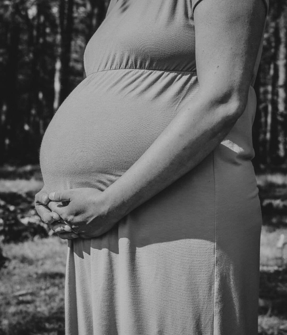 Artistieke zwart-wit foto van een zwangere vrouw, met de nadruk op haar silhouet en de ronding van haar buik, staand in een natuurlijk landschap.