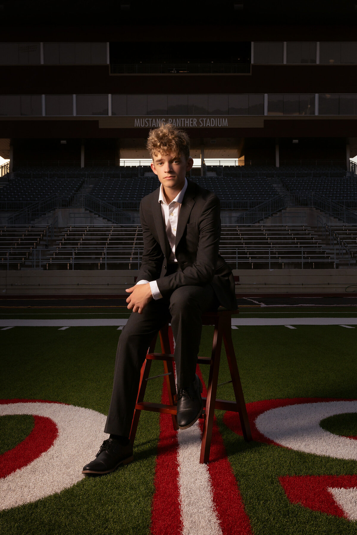 A young man in a suit sits confidently on a wooden stool on the football field of Mustang Panther Stadium, with dramatic lighting highlighting his serious expression.