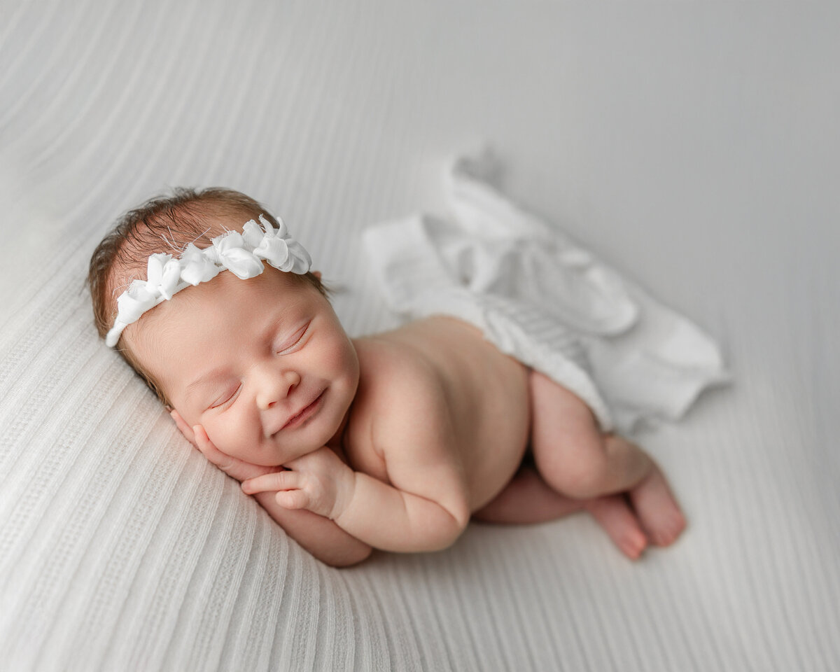 Joyful newborn portrait featuring a sleeping baby girl with a natural smile, laying on a textured white blanket. Captured by Inland Empire newborn photographer Kaitlyn Dawn.