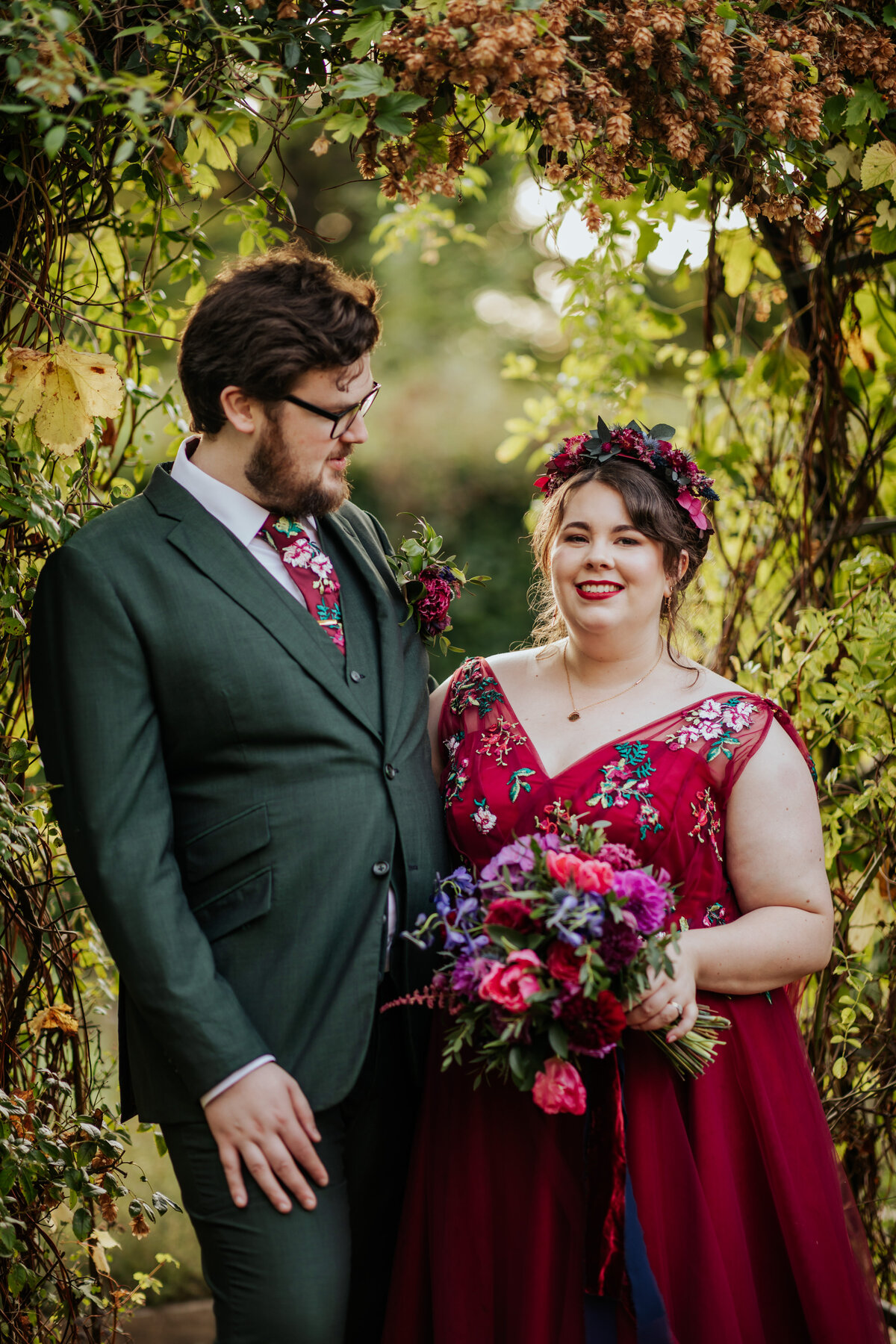 Bride wearing red wedding dress and groom in green suit stand under the arch at Ufton Court during golden hour